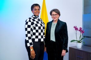 Fotografía de dos mujeres de pie en una oficina. A la izquierda, una mujer afrodescendiente con chaqueta de cuadros blancos y negros y pantalón oscuro. A la derecha, una mujer con gafas rojas y un traje de sastre oscuro. Detrás de ellas está la bandera de Colombia. A la derecha hay una orquídea morada sobre un mueble de madera. El fondo es blanco con una luz azulada tenue a la izquierda.