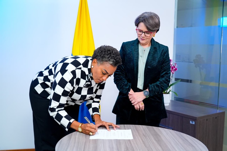 Una mujer afrodescendiente de cabello corto y chaqueta a cuadros blancos y negros está inclinada sobre una mesa redonda de madera, firmando un documento con un bolígrafo. Detrás de ella, otra mujer de cabello corto con gafas y chaqueta verde oscuro observa la firma con las manos entrelazadas. Al fondo se ve la bandera de Colombia (amarillo, azul y rojo) y una orquídea en una maceta sobre un mueble de oficina.