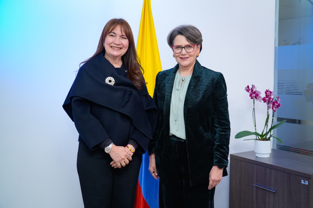 Dos mujeres de pie sonríen a la cámara en una oficina. A la izquierda, una mujer de cabello largo castaño viste capa o abrigo azul oscuro con broche plateado y pantalón negro. A la derecha, una mujer de cabello corto con gafas de marco rojo luce un traje sastre verde oscuro con blusa clara. Entre ellas está la bandera de Colombia y, al lado, una orquídea morada en maceta blanca.