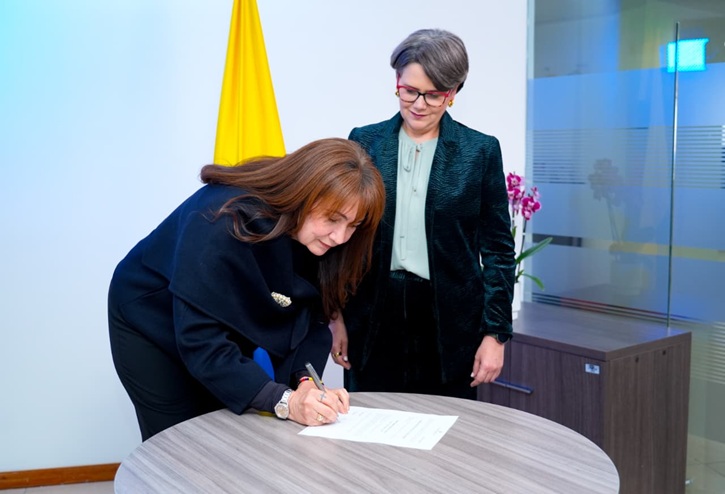 Una mujer de cabello largo castaño y abrigo azul oscuro está inclinada sobre una mesa redonda de madera, firmando un documento con bolígrafo. A su lado, una mujer de cabello corto con gafas y chaqueta verde oscuro observa el acto atenta. Al fondo se aprecia la bandera de Colombia y una orquídea morada en una maceta sobre un mueble de oficina.