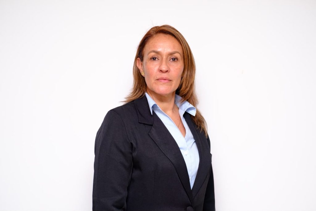 GERENTE REGIONAL CALDAS: Retrato de estudio a medio cuerpo de una mujer con cabello castaño claro y liso, peinado hacia atrás. Viste un traje de chaqueta azul marino sobre una blusa azul claro con cuello. La mujer mira directamente a la cámara con una expresión neutra. El fondo es de color blanco sólido.
