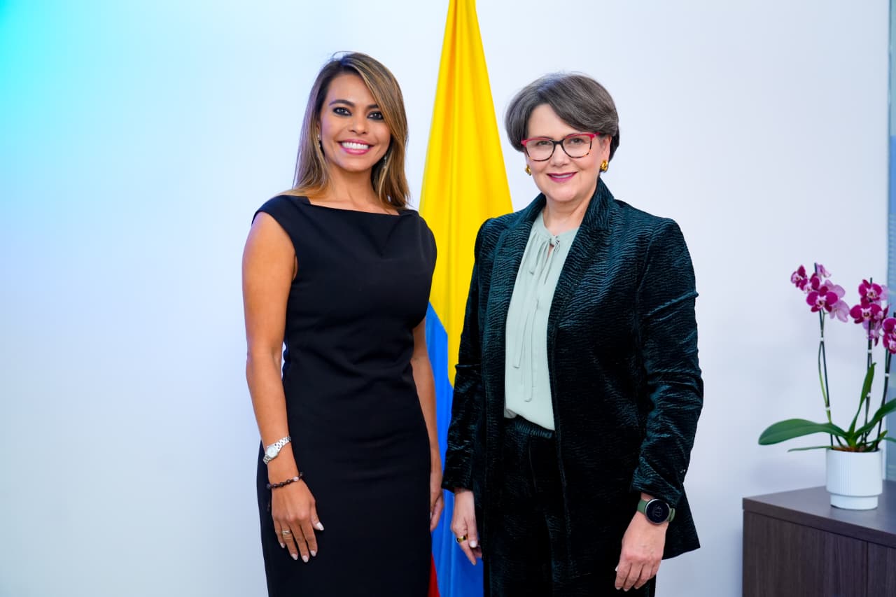 Dos mujeres posan juntas sonriendo de pie en una oficina, con la bandera de Colombia al fondo. La mujer de la izquierda lleva un vestido negro sin mangas y tiene el cabello largo castaño con reflejos claros. A la derecha, la mujer viste un saco oscuro de textura sobre una blusa clara y lleva gafas. A un costado se observa una orquídea morada en su maceta.