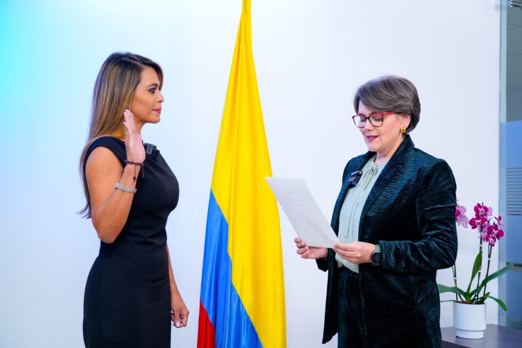 Dos mujeres participan en una ceremonia oficial frente a la bandera de Colombia. A la izquierda, una mujer con vestido negro sin mangas levanta su mano derecha en gesto de juramento. A la derecha, la mujer con saco oscuro de textura y gafas lee un documento oficial. Al fondo se observa una maceta con una orquídea morada.