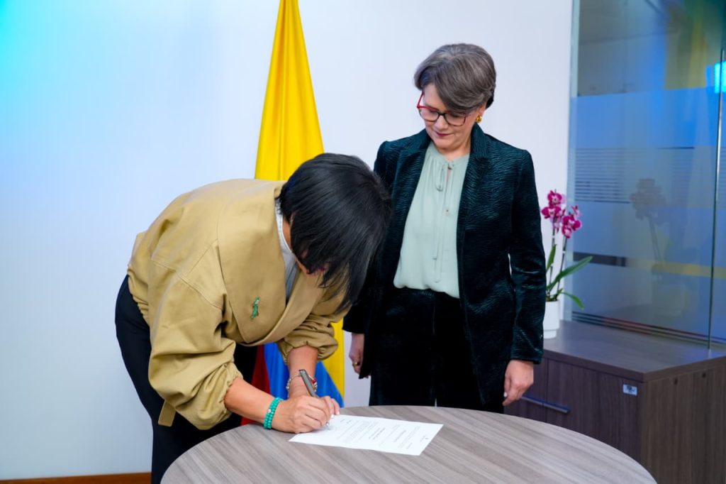 Una mujer con chaqueta beige se inclina sobre una mesa de madera para firmar un documento con un bolígrafo. A su lado, de pie, otra mujer con saco oscuro de textura y gafas observa atentamente la firma. De fondo se encuentra la bandera de Colombia y, a un costado, una maceta con una orquídea morada.
