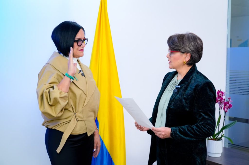 Dos mujeres se encuentran frente a frente durante un acto oficial. La mujer de la izquierda, vestida con chaqueta beige, levanta su mano derecha en gesto de juramento. Frente a ella, la mujer de saco oscuro sostiene un documento en sus manos mientras la observa. Al fondo se aprecia la bandera de Colombia y una orquídea morada a la derecha.