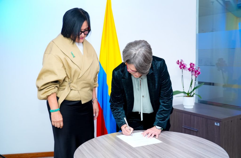 Una mujer de saco oscuro con textura y gafas de marco rojo se inclina sobre una mesa redonda para firmar un documento. A su lado, de pie, la mujer de chaqueta beige observa el momento atentamente. Al fondo destaca la bandera de Colombia y, sobre un mueble a la derecha, una orquídea morada en su maceta.