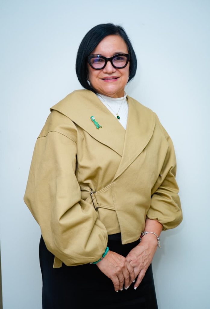 Retrato de una mujer con cabello corto negro y gafas oscuras, sonriendo hacia la cámara sobre un fondo blanco. Viste una chaqueta beige de estilo cruzado con cuello amplio y mangas abullonadas, un top blanco de cuello alto debajo y un broche verde en la solapa. Tiene las manos entrecruzadas al frente y lleva una pulsera turquesa.