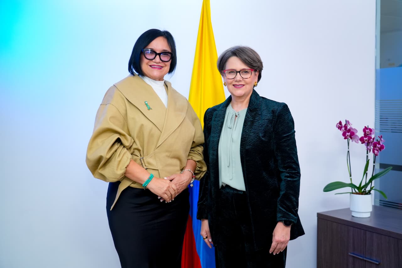 Dos mujeres posan juntas sonriendo de pie en una oficina, frente a la bandera de Colombia. La mujer de la izquierda viste una chaqueta beige cruzada y pantalón negro. La mujer de la derecha lleva un saco con textura en tono oscuro sobre una blusa clara. Ambas usan gafas. A la derecha, en una mesa, se observa una orquídea con flores moradas.