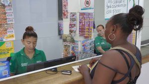 Fotografía en un punto de atención al cliente de "Gana". Una empleada con camiseta verde atiende tras un mostrador de vidrio a una mujer de espaldas. Al fondo se observan exhibidos múltiples billetes y boletos de lotería.