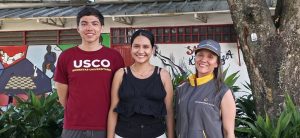 Tres personas sonríen al aire libre junto a plantas y un árbol. A la izquierda, un hombre joven con camiseta roja de la "USCO". En el centro, una mujer joven con camiseta negra de tirantes. A la derecha, una mujer con chaleco y gorra grises institucionales de "Prosperidad Social". Al fondo hay una pared pintada con murales de deportes.