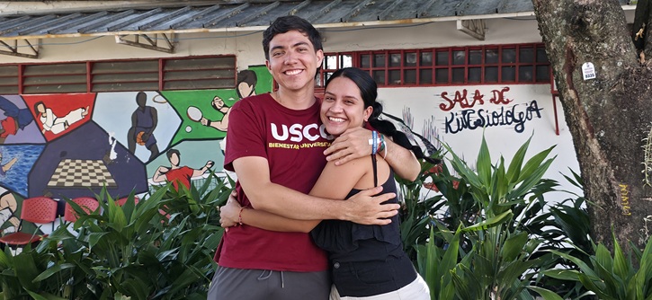 Un joven y una joven se abrazan sonrientes al aire libre, frente a un mural colorido y plantas verdes.