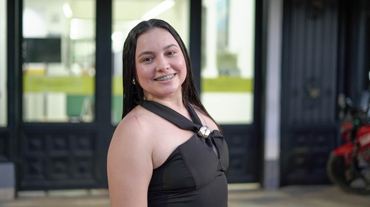 Fotografía en plano medio de una mujer joven de cabello largo y oscuro, sonriendo a la cámara y mostrando ortodoncia. Viste un elegante top negro sin mangas con un broche metálico en el escote. De fondo se aprecia una edificación urbana iluminada y parte de una motocicleta estacionada a un costado.