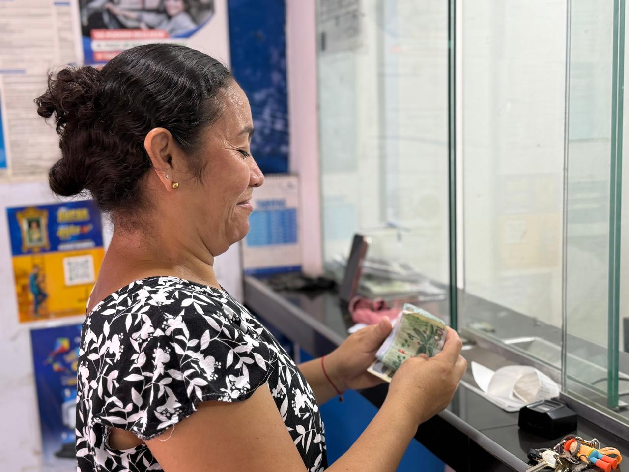 Primer plano de perfil de una mujer con blusa estampada de flores que sostiene y cuenta billetes en un punto de pago con ventanilla de vidrio.