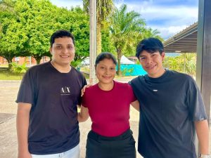 Fotografía al aire libre donde posan tres jóvenes sonrientes de pie y juntos: a la izquierda un hombre con camiseta negra con el logotipo "A|X", en el centro una mujer con blusa vinotinto y pantalón oscuro, y a la derecha un joven con camiseta negra de manga corta. De fondo se aprecian árboles frondosos y una estructura techada bajo un cielo parcialmente nublado.