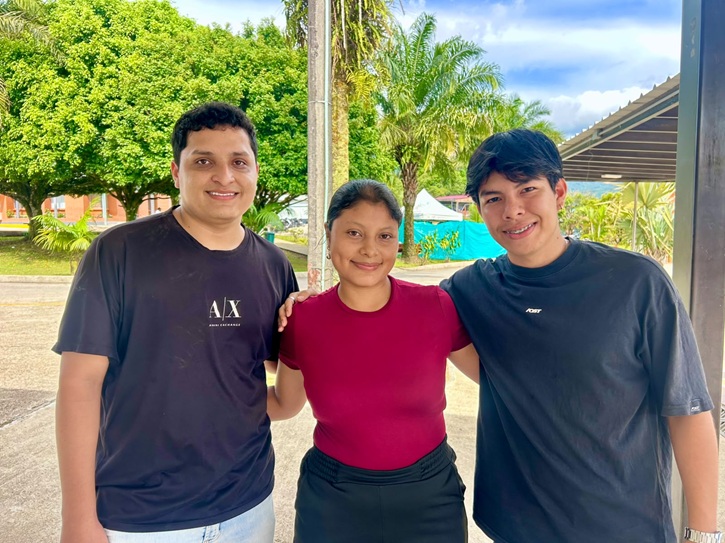 Fotografía al aire libre donde posan tres jóvenes sonrientes de pie y juntos: a la izquierda un hombre con camiseta negra con el logotipo "A|X", en el centro una mujer con blusa vinotinto y pantalón oscuro, y a la derecha un joven con camiseta negra de manga corta. De fondo se aprecian árboles frondosos y una estructura techada bajo un cielo parcialmente nublado.