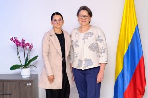 Dos mujeres posan sonrientes de pie en una oficina. A la izquierda, una mujer con gafas, cabello peinado hacia atrás, abrigo beige sobre prendas oscuras. A la derecha, una mujer con gafas de marco rojo, chaqueta beige con bordados florales y pantalón azul. A los lados destacan una orquídea morada en maceta sobre un mueble y la bandera de Colombia.