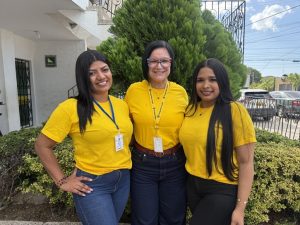 Tres mujeres sonríen de pie al aire libre frente a un arbusto y un edificio. Todas llevan camisetas amarillas. A los lados, dos mujeres jóvenes con el cabello largo y oscuro; la de la izquierda viste jeans azules y portan un carné visible. Al centro, una mujer de cabello negro corto y gafas coloridas lleva pantalones oscuros y un cordón institucional de "Prosperidad Social".
