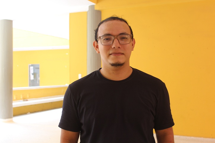 Hombre con gafas y camiseta negra de pie en un pasillo con pared de fondo amarilla.