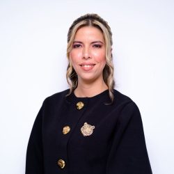 Foto Perfil Posesión Gerente Casanare Gerente Casanare: Retrato de medio cuerpo de una mujer con cabello rubio ondulado y sonriendo a la cámara. Viste una chaqueta negra con tres botones dorados grandes en la parte delantera y un prendedor dorado de la cabeza de un león en el lado izquierdo. El fondo es liso y de color blanco.