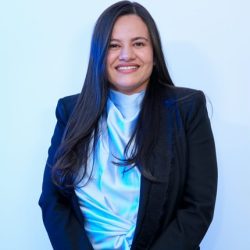Retrato formal de una mujer de plano medio, sonriendo amablemente a la cámara sobre un fondo claro con tono azulado. Tiene el cabello largo, liso y oscuro caído sobre los hombros. Viste una blusa de seda o tela satinada de color blanco o celeste claro con cuello drapeado y una elegante chaqueta o blazer negro encima. Transmite una presencia profesional y acogedora.