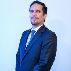 Retrato formal de un hombre de pie, visto ligeramente de perfil, sobre un fondo plano iluminado con luz azulada. Lleva un traje sastre oscuro, camisa blanca y una corbata azul brillante. Tiene cabello corto oscuro, barba de candado bien recortada y mantiene las manos cruzadas al frente con una expresión serena y profesional.