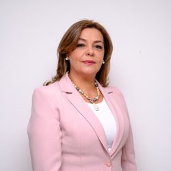 WhatsApp Image 2026-09-11 at 3.48.25 PM Fotografía de estudio a medio cuerpo de una mujer adulta sonriente sobre un fondo blanco y liso. Viste un traje de chaqueta rosa claro con solapas anchas sobre una camisa blanca. Lleva un collar de perlas de imitación con cuentas doradas, negras y grises, y pendientes de perlas redondas a juego. Su cabello oscuro y ondulado está peinado hacia atrás y peinado hacia un lado. La mujer mira directamente a la cámara con una expresión cálida y amable.