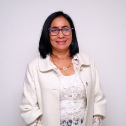 WhatsApp Image 2026-09-12 at 8.47.21 AM (3) Gerente Cundinamarca: Retrato de plano medio de una mujer de tez clara y cabello oscuro hasta los hombros. Sonríe a la cámara, usa gafas de montura morada y un collar de eslabones dorados. Viste una chaqueta blanca sobre una blusa de encaje blanco con mangas a juego. Sus manos están suavemente unidas frente a ella. El fondo es de color blanco sólido.